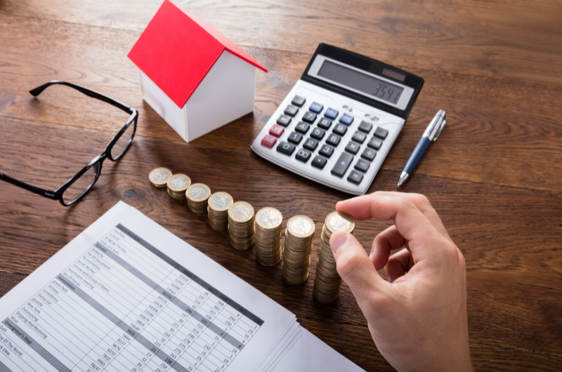 Faixas de renda do Minha Casa Minha Vida: veja qual é a sua | Foto de uma pessoa organizando moedas em uma mesa, com uma casinha, calculadora, óculos e papeis ao lado | Tudo sobre Minha Casa Minha Vida | Blog Alea