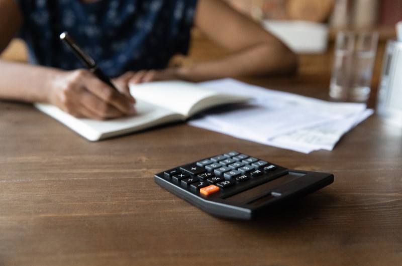 Quais são as taxas de juros de cada faixa de renda do Minha Casa Minha Vida | Foto de uma pessoa anotando em um caderno, com uma calculadora ao lado | Tudo sobre Minha Casa Minha Vida | Blog Alea