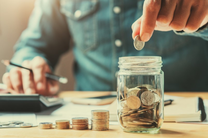 Baixas taxas de juros | Foto de um homem colocando moedas em um pote de vidro | Tudo sobre Minha Casa Minha Vida | Blog Alea
