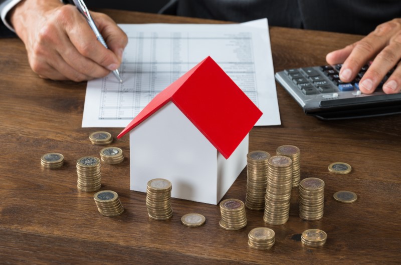 Faixas de renda do programa | Foto de uma pessoa escrevendo em um papel, com uma casinha, calculadora e moedas sobre a mesa | Tudo sobre o Minha Casa Minha Vida | Blog Alea